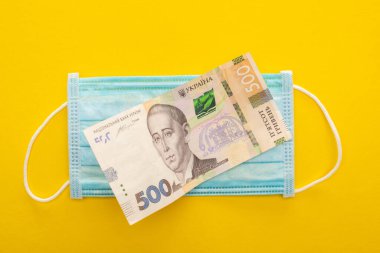 Sarı arka planda Ukrayna banknotu ve tıbbi maskesinin üst görüntüsü