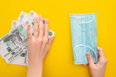 KYIV, UKRAINE - 25 Mart 2020: Rus banknotlarını kenara koyan ve sarı arka planda tıbbi maske tutan kadının kısmi görüntüsü