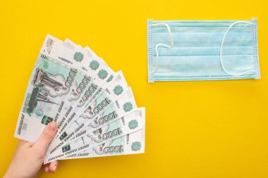 KYIV, UKRAINE - MARCH 25, 2020: partial view of woman holding Russian banknotes near medical mask on yellow background clipart