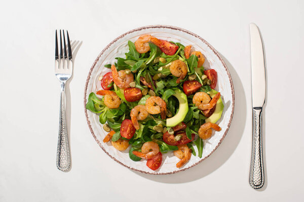 top view of fresh green salad with shrimps and avocado on plate near cutlery on white background