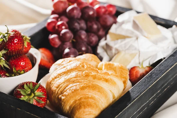 close up view of french breakfast with croissant, Camembert, grape, strawberries on wooden tray on textured white cloth