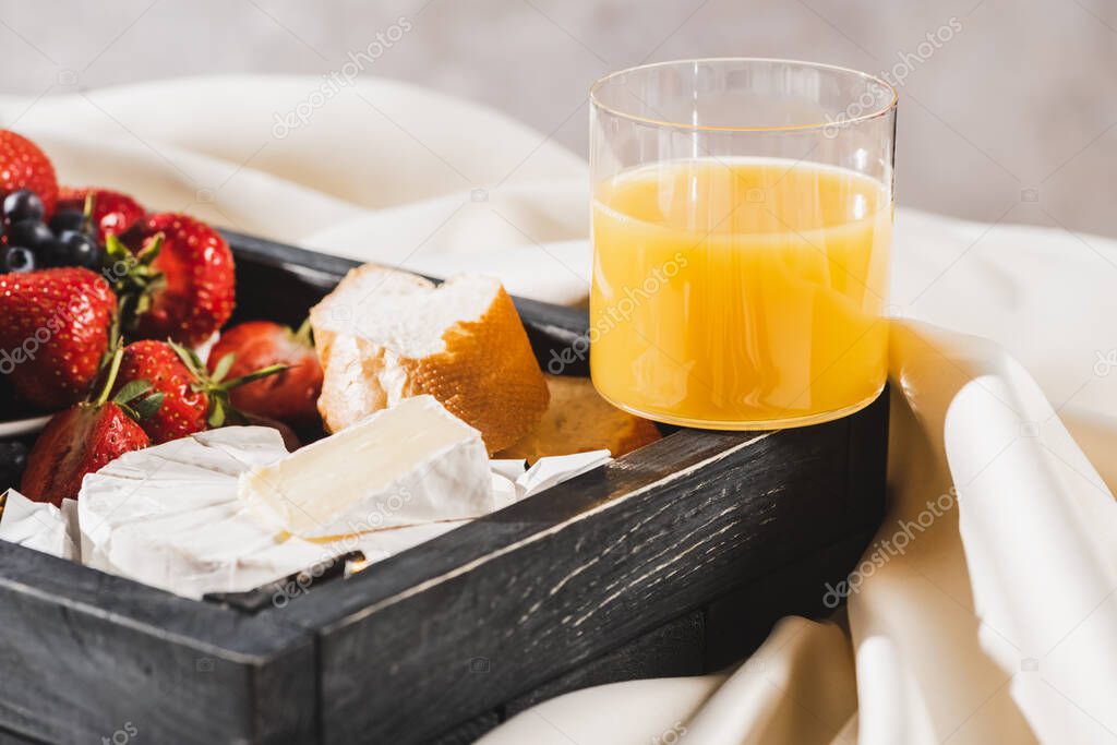 close up view of french breakfast with Camembert, orange juice, berries and baguette on wooden tray on textured white cloth