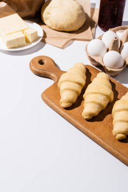 Beyaz arka planda tereyağı, yumurta ve hamurun yanındaki tahta kesme tahtasındaki çiğ kruvasanların seçici odağı