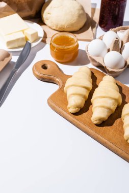 Beyaz arkaplanda reçel, tereyağı, yumurta ve hamurun yakınındaki ahşap kesme tahtasındaki çiğ kruvasanların seçici odağı