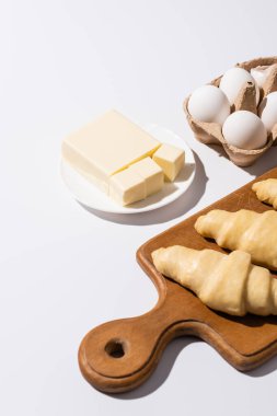 Tahta üzerinde çiğ kruvasanlar, tereyağı yanında kesim tahtası, beyaz arka planda yumurtalar.
