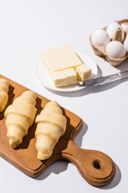 selective focus of raw croissants on wooden cutting board near butter, eggs on white background clipart