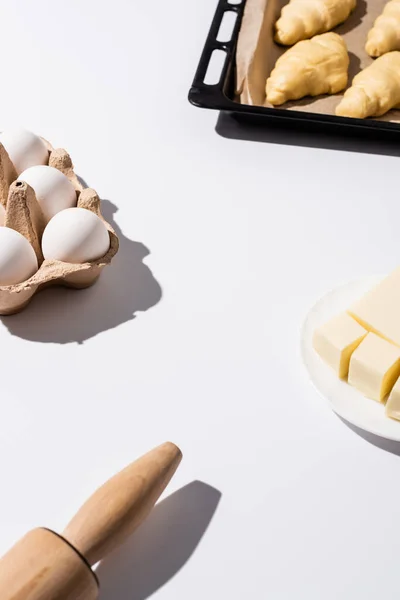 ローリングピン バター 白い背景の卵の近くのベーキングトレイに生のクロワッサンの選択的な焦点 — ストック写真