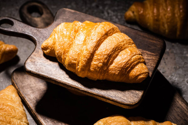 selective focus of fresh baked croissant on wooden cutting board on concrete grey surface in dark