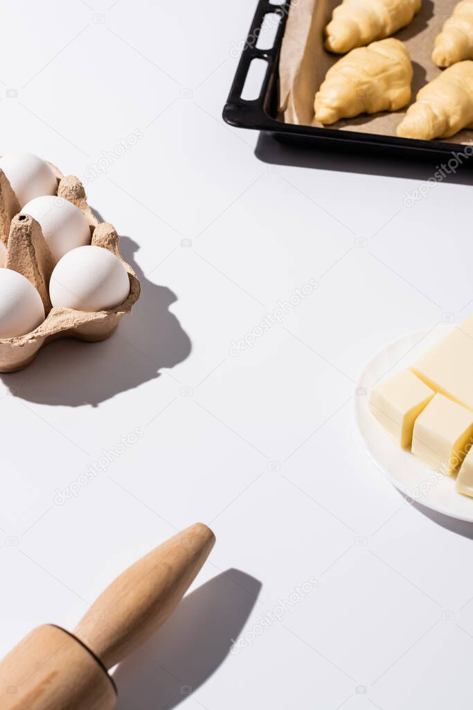 selective focus of raw croissants on baking tray near rolling pin, butter, eggs on white background