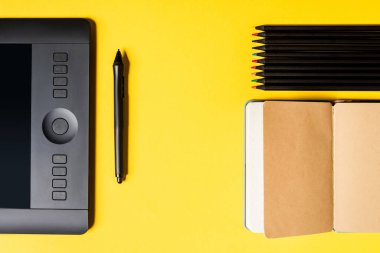 Top view of graphics tablet and stylus near open notebook and color pencils on yellow surface clipart