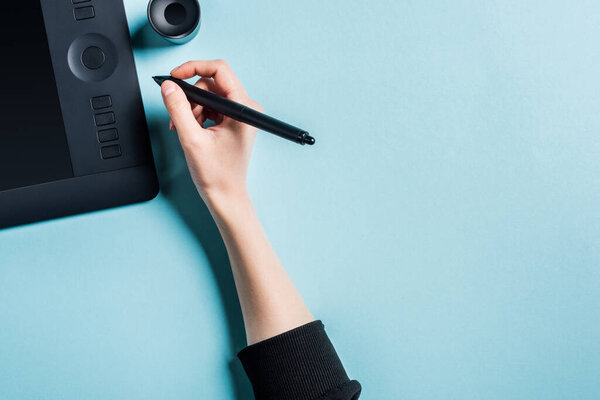 Top view of designer holding stylus near graphics tablet on blue background