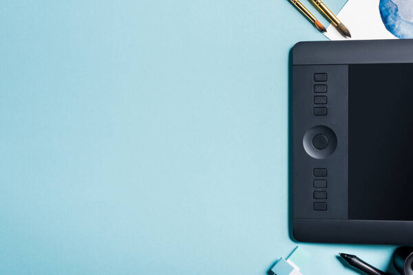 Top view of graphics tablet near paintbrushes and watercolor painting on blue background