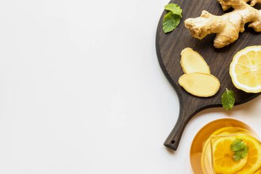 Çaydanlıktaki sıcak çayın beyaz arka planda zencefil kökü, limon ve nane ile ahşap tahtanın yanında servis edildiği üst manzara