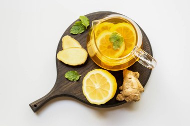Çaydanlıktaki sıcak çayın beyaz arka planda zencefil kökü, limon ve nane ile servis edilen ahşap tahtada görüntüsü.