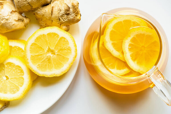 top view of hot tea in teapot near ginger root, lemon on plate on white background