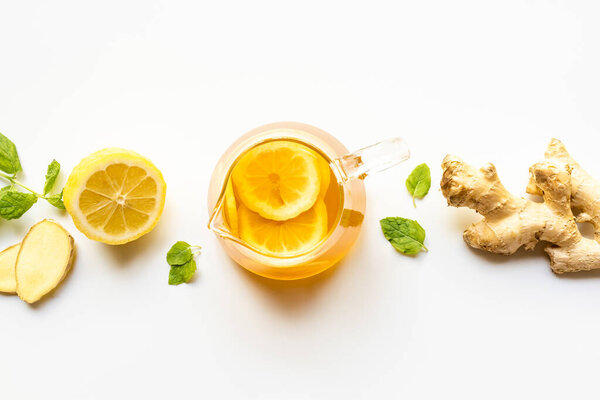 вид на горячий чай в стеклянном чайнике около корня имбиря, лимона и мяты на белом фоне
