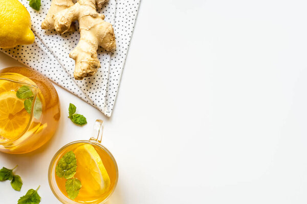 вид на горячий чай возле корня имбиря, лимона и мяты на салфетке на белом фоне
