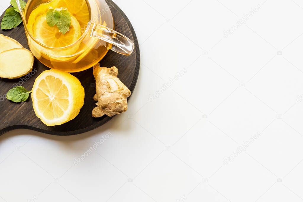 top view of hot tea in teapot served on wooden board with ginger root, lemon and mint on white background