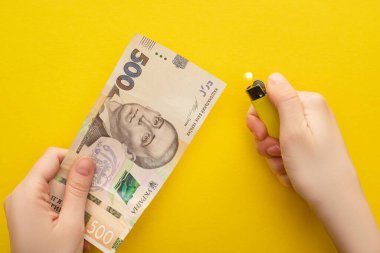 KYIV, UKRAINE - MARCH 25, 2020: cropped view of woman holding lighter near ukrainian hryvnia banknote isolated on yellow  clipart