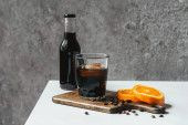 Kalter Brühkaffee mit Eis im Glas und Flasche in der Nähe von Orangenscheiben auf Schneidebrett und Kaffeebohnen auf weißem Tisch