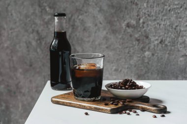 Bardakta buzlu soğuk kahve ve beyaz masanın üzerinde kahve çekirdeğinin yanında kahve şişesi.