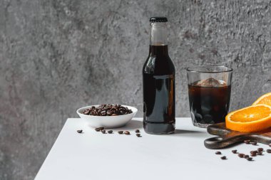 Buzlu soğuk demlenmiş kahve ve portakal dilimlerinin yanında şişe ve beyaz masada kahve çekirdekleri.