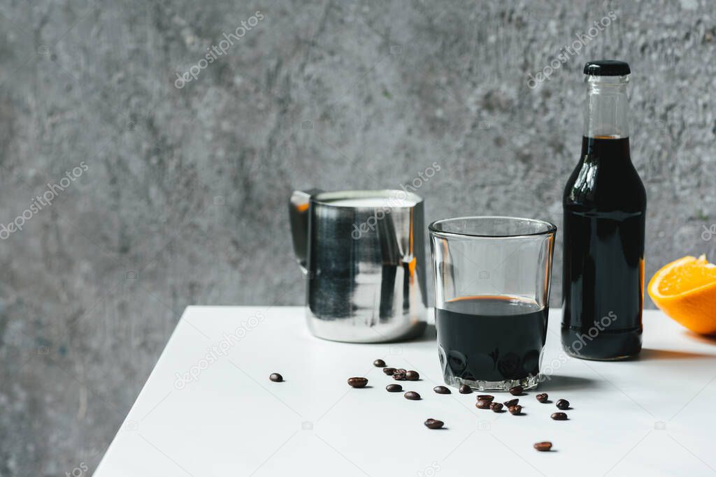 selective focus of cold brew coffee with ice in glass and bottle near orange and coffee beans on white table