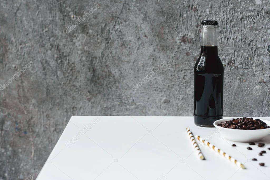 selective focus of cold brew coffee with ice in bottle near drinking straws and coffee beans on white table