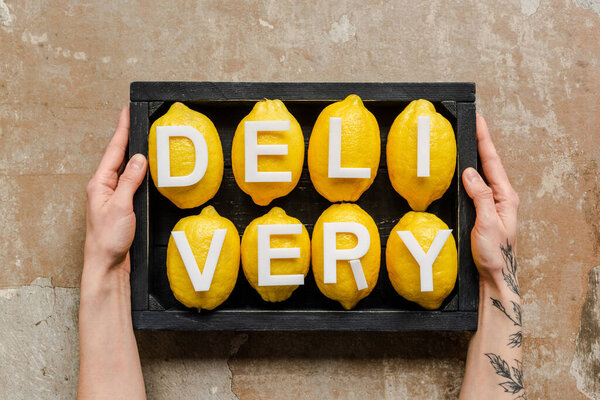partial view of woman holding wooden box with word delivery on lemons on weathered surface