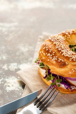 Etli taze çörek, kırmızı soğan, krem peynir ve peçetede lahanalar dokulu yüzeyde çatal bıçak takımı.