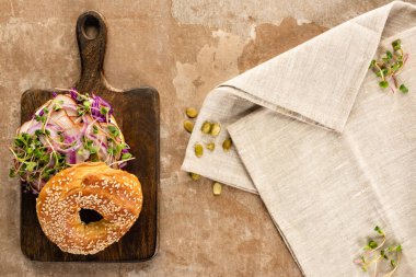top view of fresh delicious bagel with meat, red onion and sprouts on wooden cutting board near napkin on aged beige surface clipart