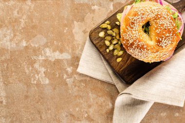 top view of fresh delicious bagel on wooden cutting board with pumpkin seeds on aged beige surface with napkin clipart