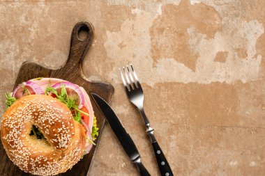 top view of fresh delicious bagel on wooden cutting board on aged beige surface with cutlery clipart