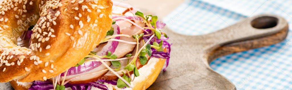 close up view of fresh delicious bagel with meat, red onion, cream cheese and sprouts on wooden cutting board near napkin, panoramic shot