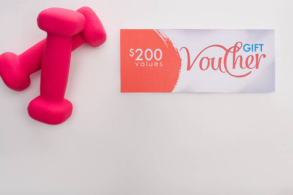Вид сверху розовых гантелей рядом с подарочным ваучером стоимостью 200 долларов на белом фоне
