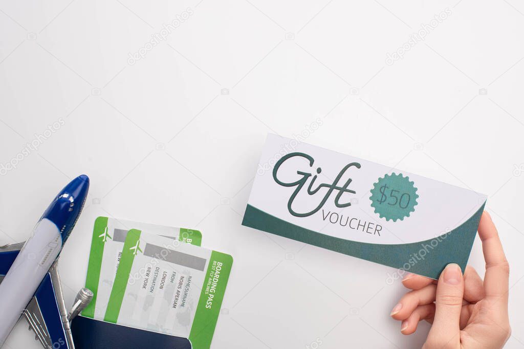 Top view of woman holding gift voucher near passport with air tickets and toy airplane on white background