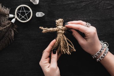 top view of woman holding voodoo doll in hands near pentagram and crystals on black  clipart