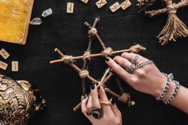 top view of witch touching pentagram near runes, book, crystals, skull and voodoo doll on black  clipart