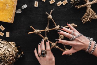 cropped view of witch touching pentagram near runes, book, crystals, skull and voodoo doll on black  clipart