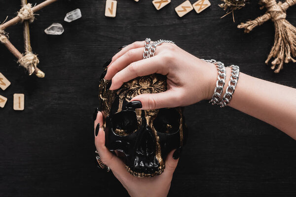 cropped view of woman holding skull near crystals and runes on black 