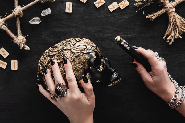 cropped view of woman holding candle near skull, runes and crystals on black 