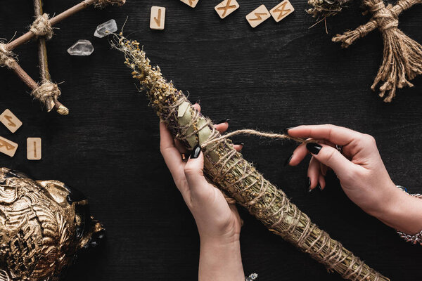 top view of woman holding dried herbs near runes, voodoo doll, runes, skull and crystals on black 