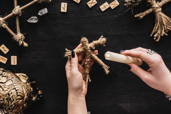 top view of woman holding burning candle near voodoo doll, runes, skull and crystals on black 