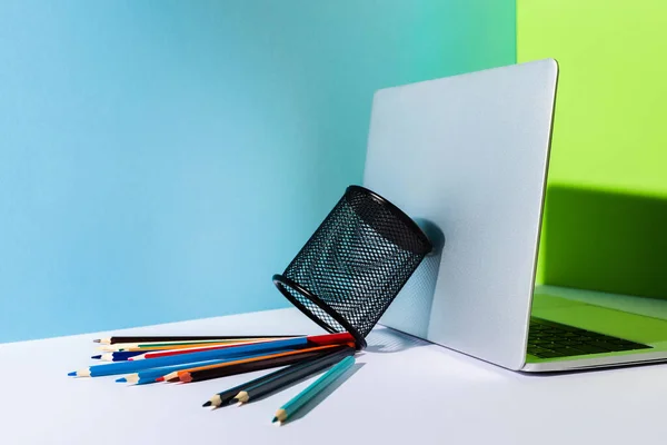 青緑白の背景に現代のノートパソコンの近くにある鉛筆ホルダーから色鉛筆を撒き散らし — ストック写真