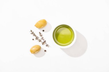 top view of olive oil in bowl near green olives, herb and black pepper on white background clipart
