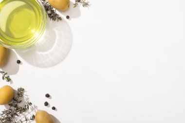 Yeşil zeytinlerin yanındaki cam kasede zeytinyağı, bitkiler ve beyaz arka planda siyah biber manzarası.