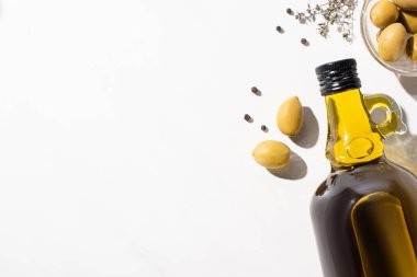Şişede zeytinyağının en üst görüntüsü, yeşil zeytin kabının yanında, otlar ve siyah biberler beyaz arka planda.