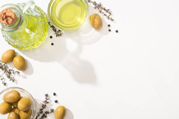 top view of olive oil in bowl and bottle near green olives, herb and black pepper on white background