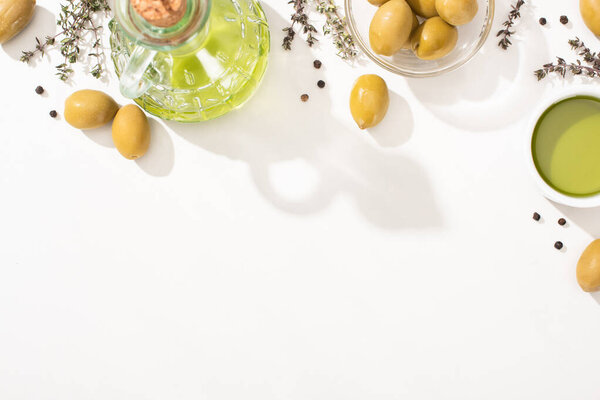 top view of olive oil in glass bowl and bottle near green olives, herb and black pepper on white background