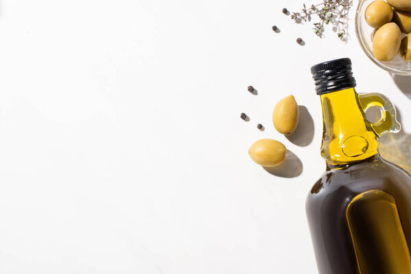 top view of olive oil in bottle near green olives in bowl, herb and black pepper on white background